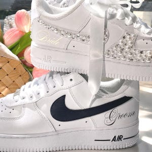 May include: A pair of white sneakers. The top shoe features white satin laces, pearl and rhinestone embellishments, and the word Bride in script. The bottom shoe features a black Nike swoosh logo and the word Groom in script on the side.