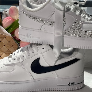May include: Two white sneakers stacked vertically. The top sneaker features white satin ribbon laces and is embellished with white pearls and clear rhinestones along the side panel. The bottom sneaker has a black Nike swoosh and white laces.