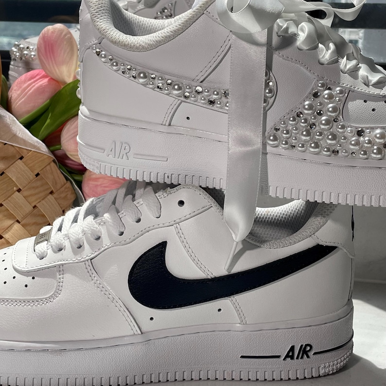 May include: Two white sneakers stacked vertically. The top sneaker features white satin ribbon laces and is embellished with white pearls and clear rhinestones along the side panel. The bottom sneaker has a black Nike swoosh and white laces.