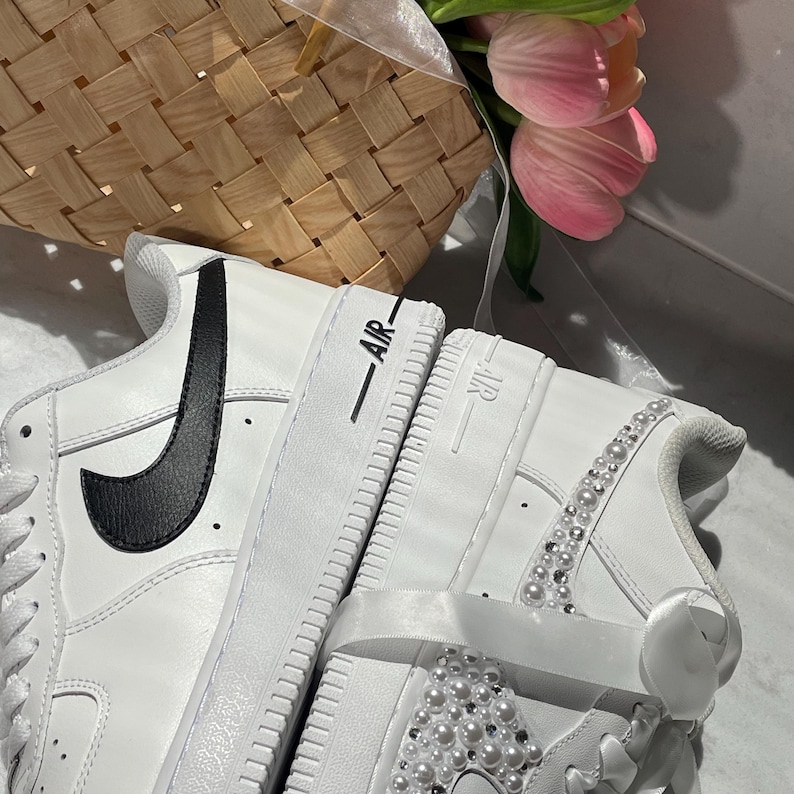 May include: A pair of white sneakers featuring a black swoosh logo on one side and white pearl embellishments on the other. The midsoles are embossed with the text AIR. The shoes include white satin ribbon laces and are positioned near pink tulips.