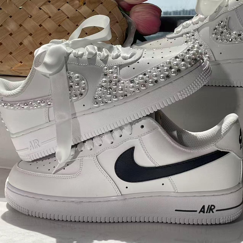 May include: A pair of white sneakers featuring a low-top silhouette with a thick rubber sole. One sneaker is embellished with numerous white pearls along the side and toe box, finished with wide white satin ribbon laces. The other sneaker has a black swoosh.