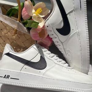 May include: A pair of white low-top sneakers featuring a black Nike swoosh logo on the side and the word AIR embossed on the white rubber midsole. The sneakers have white laces, a perforated toe box, and a smooth leather-like upper construction.