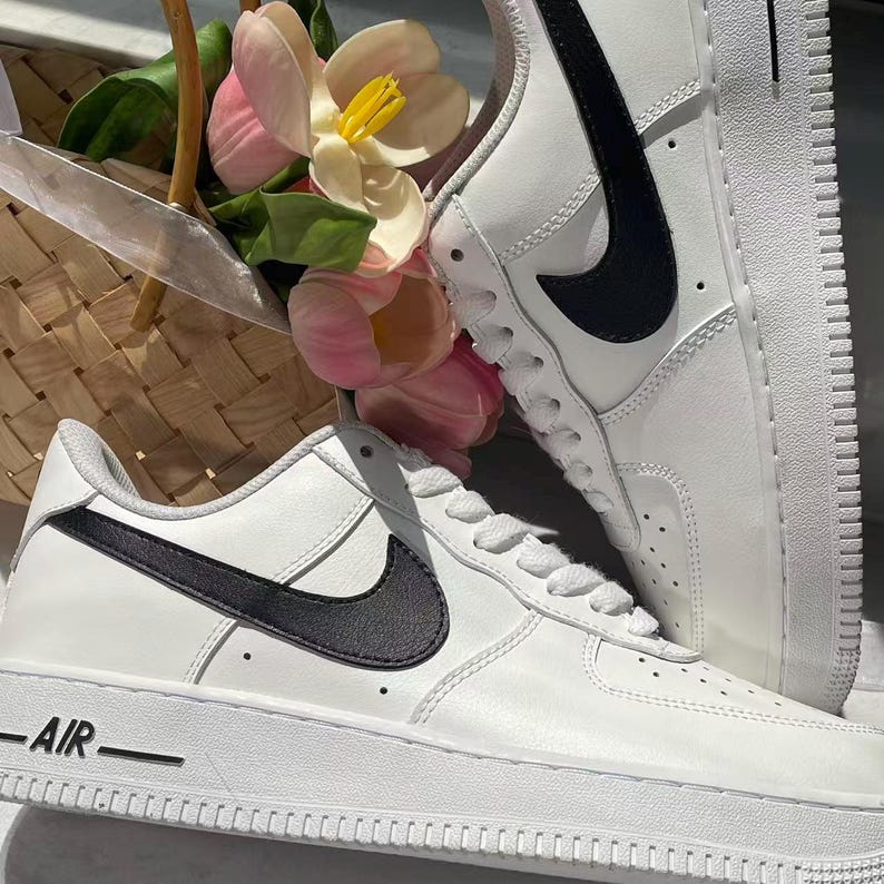 May include: A pair of white low-top sneakers featuring a black Nike swoosh logo on the side and the word AIR embossed on the white rubber midsole. The sneakers have white laces, a perforated toe box, and a smooth leather-like upper construction.