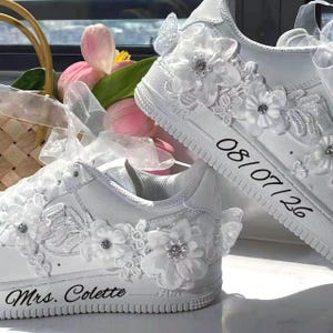 May include: A pair of white sneakers decorated with white fabric flowers, lace appliques, and small pearls. One shoe features the text 'Mrs. Colette' in black script, while the other displays the date '08/07/26' in black text on the side.