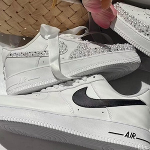 May include: Two white sneakers. The top sneaker features white satin ribbon laces and is embellished with rows of white pearls and clear rhinestones along the side. The bottom sneaker has a smooth white leather finish with a black Nike swoosh logo.