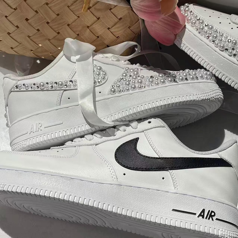 May include: Two white sneakers. The top sneaker features white satin ribbon laces and is embellished with rows of white pearls and clear rhinestones along the side. The bottom sneaker has a smooth white leather finish with a black Nike swoosh logo.