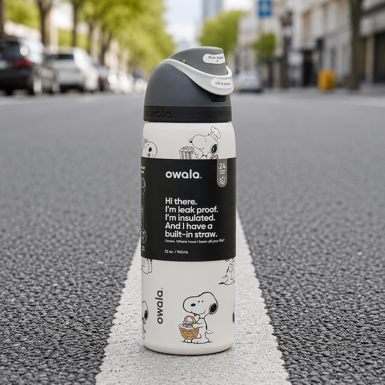 May include: A white Owala water bottle with a dark gray lid and a black label featuring Snoopy illustrations. The label text reads: Hi there. I'm leak proof. I'm insulated. And I have a built-in straw. I know. Where have I been all your life? 32 oz / 945 mL.