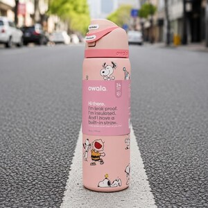 May include: A light pink Owala insulated water bottle featuring a Peanuts character print with Snoopy and Charlie Brown. The bottle has a push-button lid with a locking mechanism, a built-in straw, and a label stating 24 ounces and 710 milliliters.