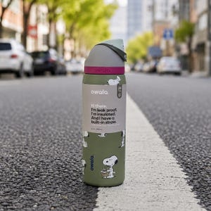 May include: An olive green Owala insulated water bottle featuring a Snoopy graphic and a gray lid with a pink accent. The bottle includes a label with the text: Hi there. I'm leak proof. I'm insulated. And I have a built-in straw. It holds 32 oz.