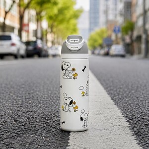 May include: A white cylindrical water bottle featuring a repeating pattern of Snoopy and Woodstock characters with small black bones. The bottle has a grey lid with a push button and a clip to unlock mechanism clearly printed on the top section.