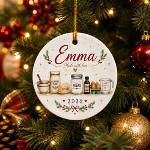 Peut inclure: Une décoration ronde en céramique blanche portant le prénom Emma, le texte Made with love et l'année 2026. Le motif illustre un bol, de la farine, du beurre, du sucre, de l'extrait de vanille, des œufs et un verre doseur avec des détails dorés.