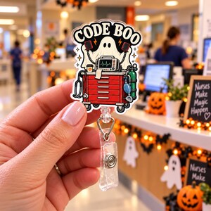 May include: An acrylic badge reel featuring a cartoon ghost standing behind a red medical crash cart with the text CODE BOO. The cart includes a heart monitor, a stethoscope, a coffee cup, and an oxygen tank. The design is attached to a clear plastic clip.