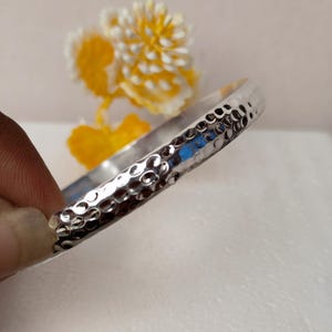 Könnte beinhalten: Ein silberfarbener Armreif mit einer gehämmerten Textur auf der gesamten Außenfläche. Das Metallband hat eine reflektierende Oberfläche mit zahlreichen kleinen, kreisförmigen Vertiefungen, die ein genopptes Erscheinungsbild erzeugen.
