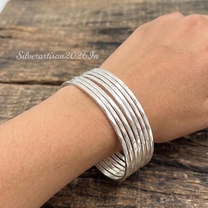 May include: A set of seven individual silver bangles featuring a hammered, textured finish. Each bangle has a reflective, metallic surface and a rounded profile. The text Silverartisan2026In is visible in the upper left corner of the image.
