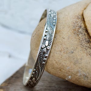 Può includere: Un bracciale rigido in argento con finitura ossidata, caratterizzato da intricati motivi floreali in rilievo e delicati dettagli a perline. La superficie metallica presenta uno sfondo testurizzato e scuro che mette in risalto i motivi floreali in argento lucido.