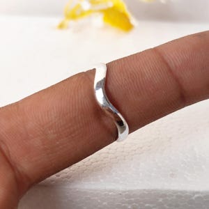 Puede incluir: Un anillo de plata pulida con un diseño de banda ondulada o serpentina continua y lisa. La superficie metálica es reflectante y presenta una forma sencilla y fluida que se curva suavemente alrededor del dedo, mostrando un acabado uniforme.