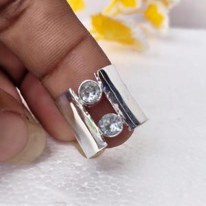 Peut inclure: Une bague en argent composée de deux bandes verticales parallèles reliées par un pont central, ornée de deux pierres rondes, claires et facettées, placées l'une au-dessus de l'autre entre les sections en argent, créant un design ajouré.