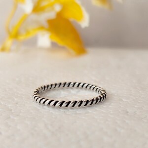 Puede incluir: Un anillo fino de plata con un diseño continuo de cuerda trenzada. El metal tiene un acabado pulido con zonas hundidas oscurecidas entre los segmentos trenzados, creando una textura distintiva que rodea toda la circunferencia del aro.