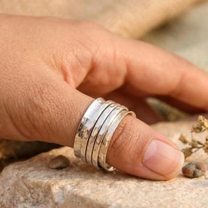 May include: A wide silver ring featuring a multi-band design with a hammered, textured finish. The ring consists of several interconnected silver bands that wrap around the finger, creating a layered appearance with a polished, reflective surface.
