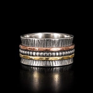 Può includere: Un anello a fascia larga con un filo d'argento perlato centrale, affiancato da una fascia in rame martellato e una in ottone martellato, inserito tra due fasce esterne in argento con texture spazzolata verticale e finitura ossidata.