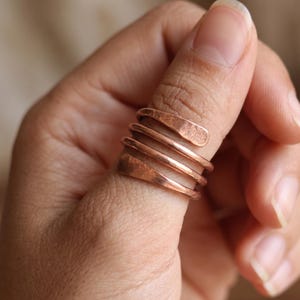 Può includere: Un anello color rame caratterizzato da un design a spirale che si avvolge più volte attorno al dito. La fascia metallica presenta una finitura martellata e testurizzata alle estremità, creando un aspetto distintivo dal tono bruno-rossastro.