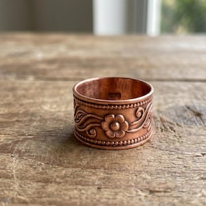 Puede incluir: Un anillo ancho de cobre que presenta un motivo floral central en relieve rodeado de patrones de enredaderas. La banda está bordeada por dos filas de pequeños detalles de cuentas en relieve a lo largo de los bordes superior e inferior, con una superficie interior lisa.