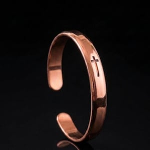 May include: A polished copper-toned cuff bracelet featuring a smooth, rounded band with an open design. A small, dark, vertical cross symbol is engraved into the surface of the metal, positioned towards the upper curve of the bracelet.