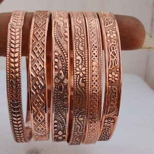 Pode incluir: Um conjunto de seis pulseiras de cobre com padrões em relevo distintos, incluindo motivos de folhas, formas geométricas de diamante, desenhos florais e volutas intrincadas. Cada pulseira tem um acabamento metálico polido.
