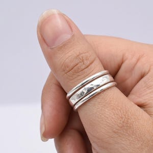 May include: A set of three silver-colored rings worn on a thumb. The outer two rings feature a smooth, polished finish, while the central ring has a textured, hammered surface. The rings are stacked together, creating a layered appearance on the finger.