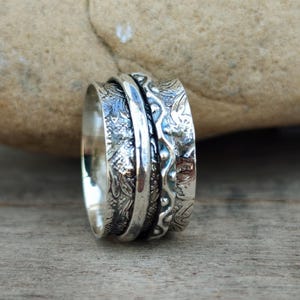 Può includere: Un anello spinner in argento caratterizzato da una fascia larga con finitura ossidata e complessi intagli floreali. Il centro dell'anello include una fascia in argento lucido e liscia, affiancata da due sezioni testurizzate con motivi in rilievo.