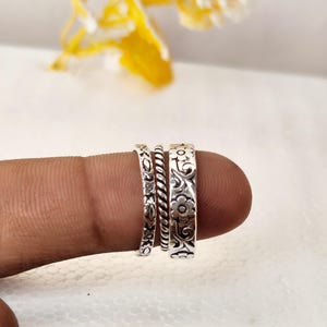 May include: A set of three silver-toned rings featuring distinct textures. One ring has a floral pattern, the second is a twisted rope design, and the third displays a wider band with an intricate embossed floral and vine motif across the surface.