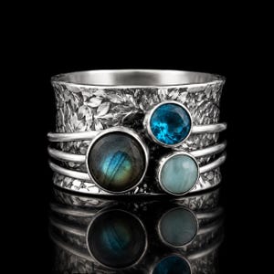 May include: A wide silver spinner ring featuring a textured leaf pattern on the band. The ring is adorned with three round gemstones: a large iridescent labradorite, a faceted blue topaz, and a smooth, pale blue cabochon stone.