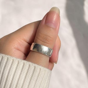 Puede incluir: Un anillo de banda ancha de plata pulida con una superficie lisa y plana y un acabado metálico de alto brillo. El anillo presenta un diseño sencillo y sin adornos con bordes rectos y limpios, reflejando la luz en su exterior plateado sólido.