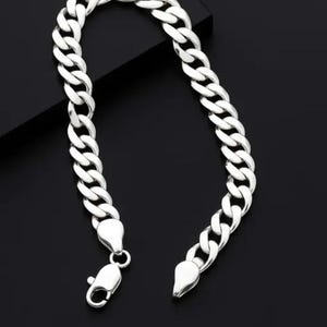 May include: A silver-toned curb chain bracelet featuring interlocking flat, oval-shaped links. The bracelet is finished with a polished metallic surface and includes a secure lobster claw clasp mechanism at one end for fastening the piece.
