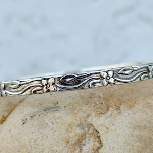 May include: A silver bangle bracelet featuring a continuous embossed pattern of small four-petaled flowers and winding leafy vines. The metal has a darkened, oxidized finish in the recessed areas to highlight the intricate floral details.