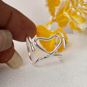 Op de afbeelding: Een zilverkleurige ring met een open, draadachtig ontwerp dat in het midden een hartvorm vormt. De band is gemaakt van één doorlopend stuk metaal dat buigt om het hartmotief te creëren, met open uiteinden aan beide zijden.