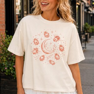 May include: An off-white short-sleeve t-shirt featuring a circular graphic print in a reddish-brown hue. The design includes a central crescent moon surrounded by multiple ladybugs and small star shapes. The shirt is paired with a white tiered mini skirt.