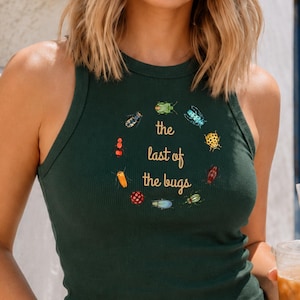 May include: A dark green ribbed tank top featuring a circular arrangement of colorful insect illustrations surrounding the gold text that reads the last of the bugs, paired with light blue denim jeans and dark-tinted round sunglasses.