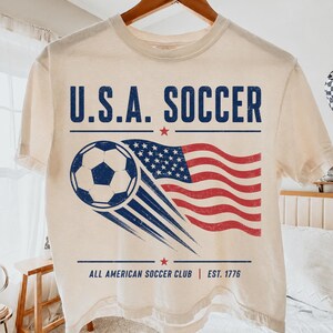 May include: A cream-colored short-sleeve t-shirt featuring a graphic with the text U.S.A. SOCCER, a soccer ball, a waving American flag, and the text ALL AMERICAN SOCCER CLUB EST. 1776 in blue and red ink with a distressed, vintage-style finish.