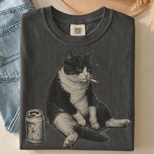 May include: A charcoal gray t-shirt featuring a graphic of a black and white cat sitting next to a silver soda can. The cat has a cigarette in its mouth. The shirt has a crew neckline and a Comfort Colors brand label printed on the inside collar.