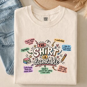 May include: A cream-colored t-shirt featuring a colorful graphic with the text "THE SHIRT is decodable" surrounded by educational linguistic notes, small stars, hearts, a stack of three books, and a pencil illustration in various pastel colors.