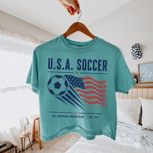 May include: A teal short-sleeve t-shirt featuring a graphic of a soccer ball and a waving American flag. The text reads U.S.A. SOCCER, ALL AMERICAN SOCCER CLUB, and EST. 1776. The design is printed in dark blue and red with a distressed texture.
