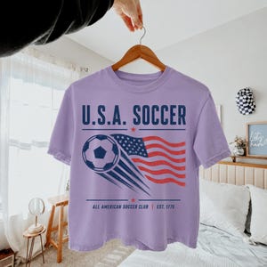 May include: A light purple t-shirt featuring a graphic with the text U.S.A. SOCCER, a soccer ball, and a waving American flag. Below the graphic, the text reads ALL AMERICAN SOCCER CLUB EST. 1776, accented by two small red stars on the top and bottom.