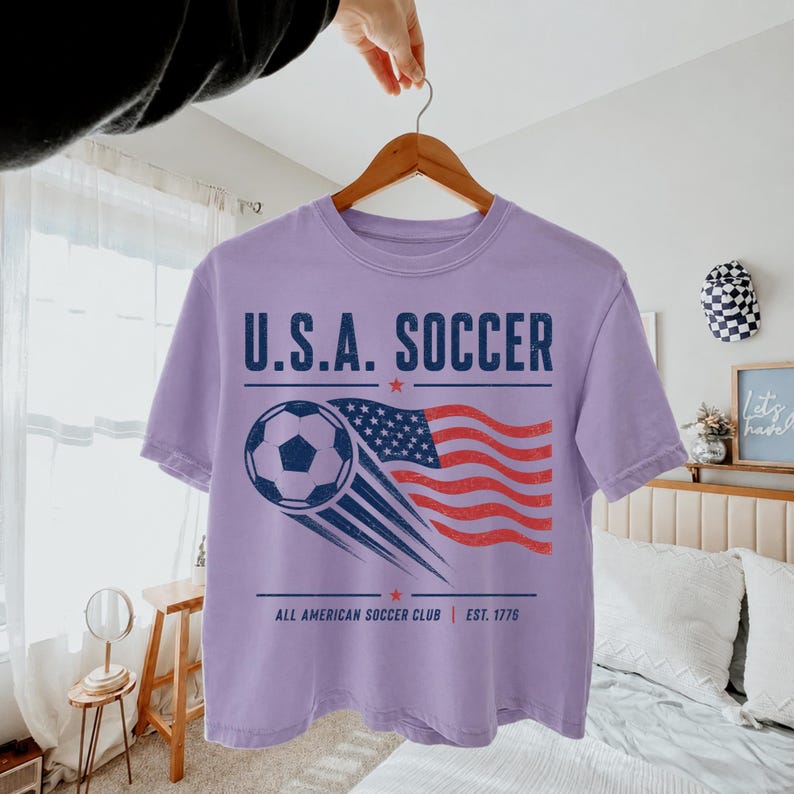 May include: A light purple t-shirt featuring a graphic with the text U.S.A. SOCCER, a soccer ball, and a waving American flag. Below the graphic, the text reads ALL AMERICAN SOCCER CLUB EST. 1776, accented by two small red stars on the top and bottom.