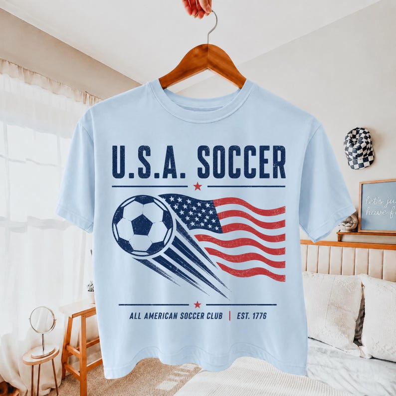 May include: A light blue t-shirt featuring a graphic of a soccer ball and a waving American flag. The text reads U.S.A. SOCCER and ALL AMERICAN SOCCER CLUB EST. 1776 in dark blue. The design has a distressed, vintage-inspired texture throughout.
