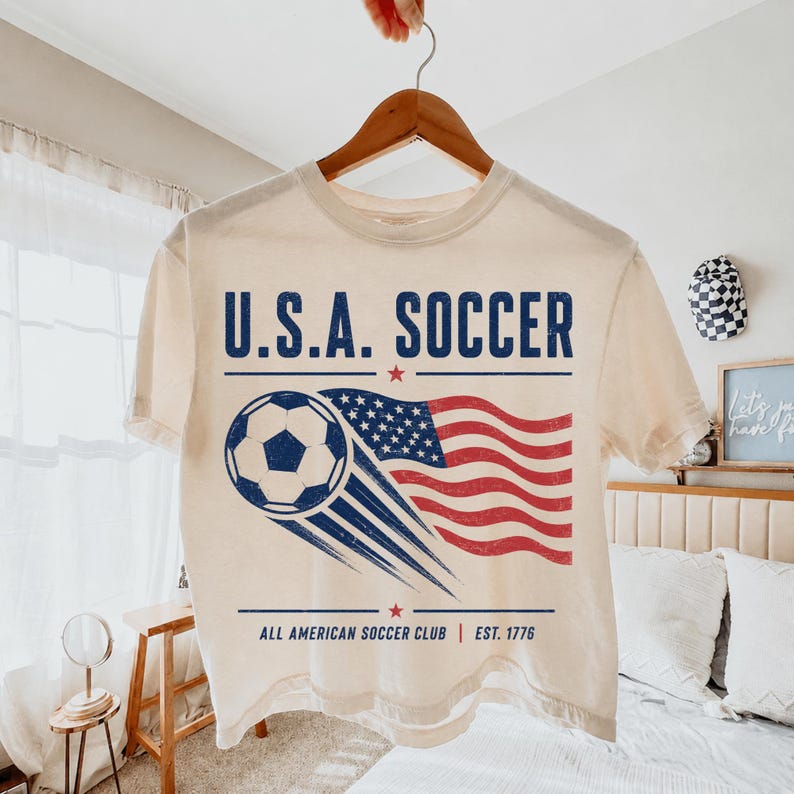 May include: A cream-colored short-sleeve t-shirt featuring a graphic with the text U.S.A. SOCCER, a soccer ball, a waving American flag, and the text ALL AMERICAN SOCCER CLUB EST. 1776 in blue and red ink with a distressed, vintage-style finish.