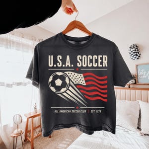 May include: A charcoal gray t-shirt featuring the text U.S.A. SOCCER in a distressed white font. Below the text is a graphic of a soccer ball with motion lines trailing into a stylized American flag. The bottom text reads ALL AMERICAN SOCCER CLUB EST. 1776.