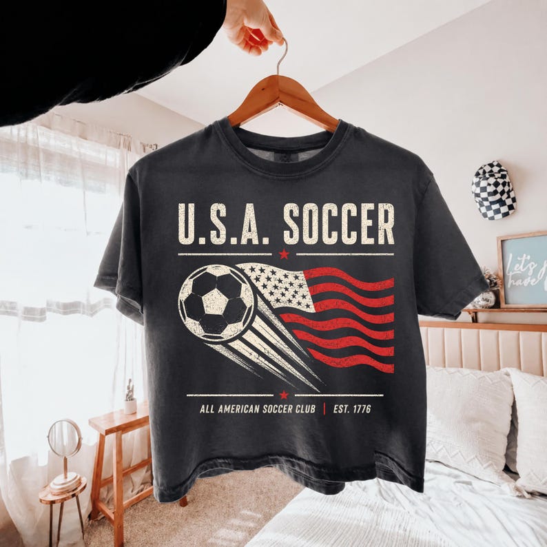May include: A charcoal gray t-shirt featuring the text U.S.A. SOCCER in a distressed white font. Below the text is a graphic of a soccer ball with motion lines trailing into a stylized American flag. The bottom text reads ALL AMERICAN SOCCER CLUB EST. 1776.