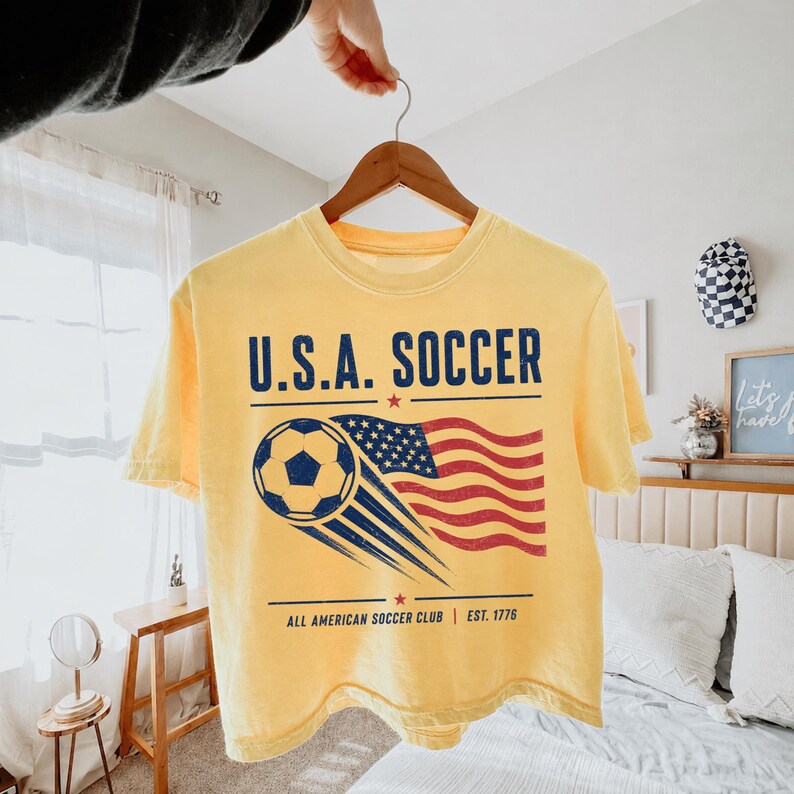 May include: A pale yellow short-sleeve t-shirt featuring a graphic print with the text U.S.A. SOCCER, a soccer ball, and a waving American flag. Below the graphic, the text reads ALL AMERICAN SOCCER CLUB | EST. 1776 with two small red stars.