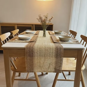 May include: A light wood dining table with four matching chairs, featuring a woven tan and brown table runner. Four beige placemats are set with white bowls. A clear glass vase with dried flowers sits in the centre under a white pendant light.